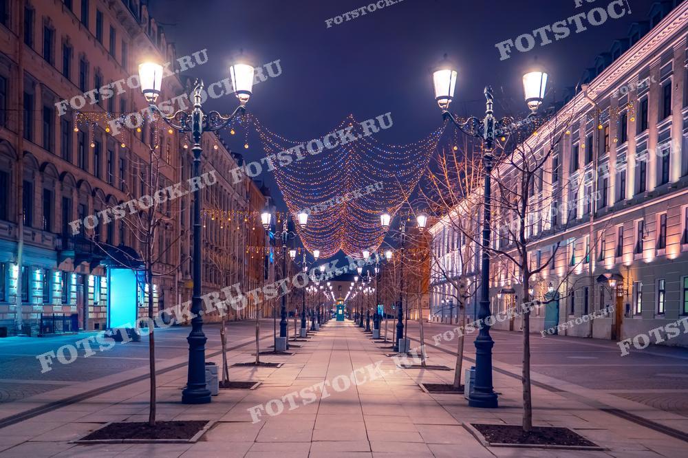 На фотографии изображена празднично украшенная улица в Санкт-Петербурге ночью. Вечерние огни и гирлянды создают атмосферу уюта и волшебства. Пустынные тротуары и освещенные здания добавляют ощущение спокойствия и тишины. Этот вид передает зимнее настроение и красоту ночного города.